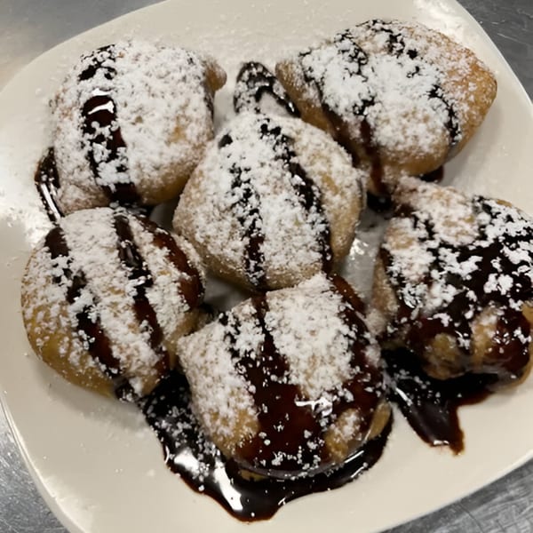 Six golden-brown fried dough balls, dusted with powdered sugar and drizzled with chocolate sauce.