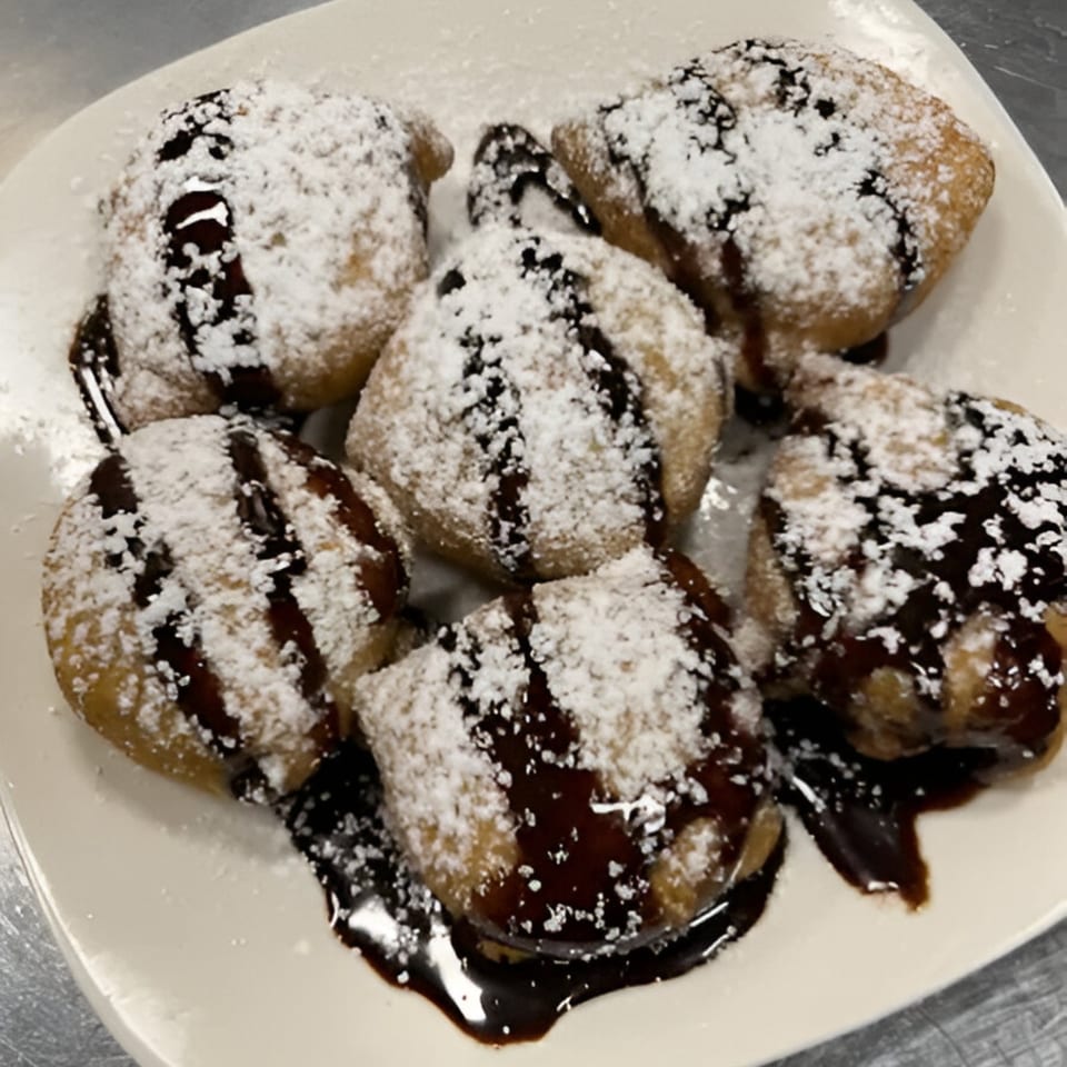 Six golden-brown fried dough balls, dusted with powdered sugar and drizzled with chocolate sauce.
